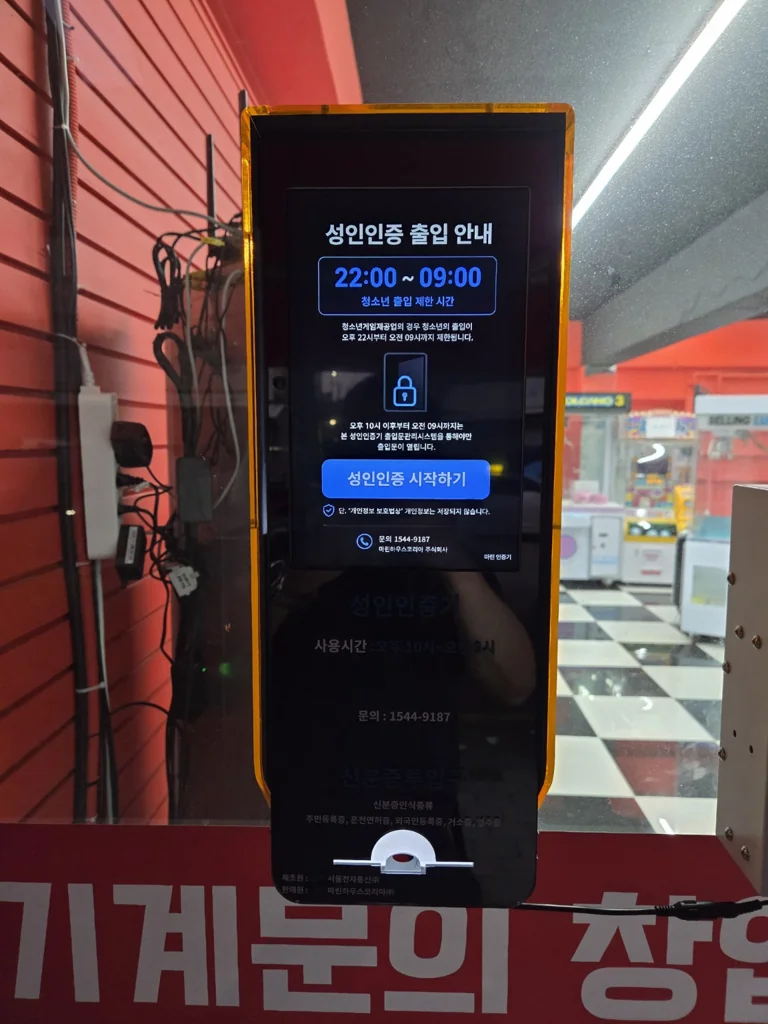 오락실 유리 출입구에 설치된 금색 성인인증기의 안내 화면과 하단 신분증 투입구